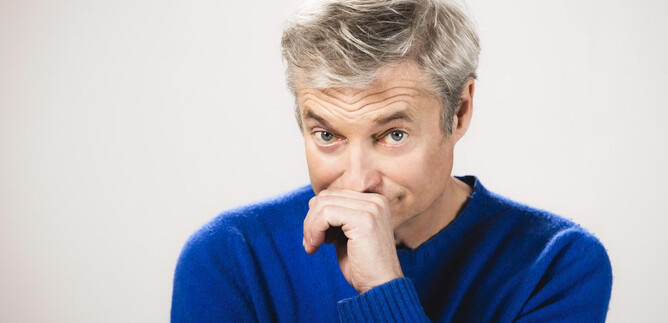 Ein Mann mit grauen Haaren trägt einen blauen Pullover. Er hat einen nachdenklichen Ausdruck und hält sich den Finger an die Lippen. | © Moritz Schell