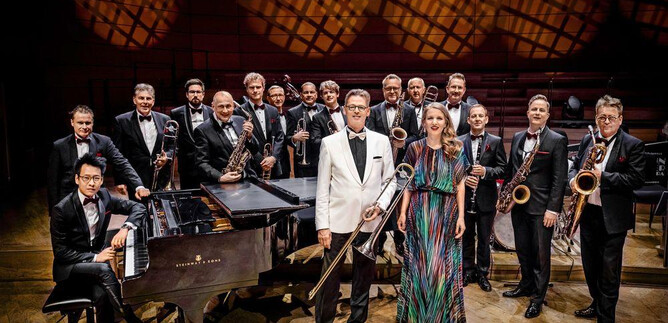 A large jazz band is on a stage, all members are dressed in formal attire. In the foreground, a piano and a singer can be seen. | © Schmidt & Salden