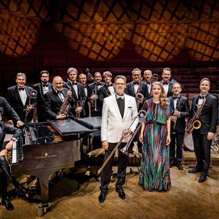 Duża orkiestra jazzowa stoi na scenie, wszyscy członkowie mają na sobie eleganckie stroje. Na pierwszym planie widać fortepian i śpiewaczkę. | © Schmidt & Salden
