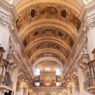 An impressive church interior with high vaults and elaborate ceiling paintings. On both sides, there are large organs, and elegant light illuminates the space. | © Erzdiözese Salzburg