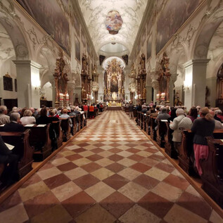 Překrásný kostel s krásnou klenbou a mnoha dekoračními detaily. Mnoho lidí sedí v lavicích a poslouchá akci. | © Erzabtei St. Peter