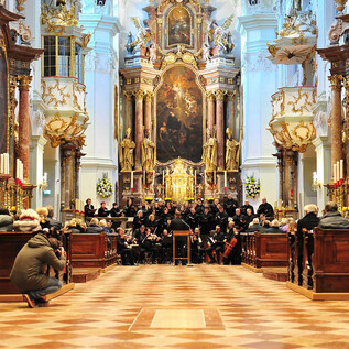 Un concert dans une église impressionnante, avec un grand groupe de musiciens et un autel artistique en arrière-plan. Les spectateurs sont assis sur des bancs en bois et profitent de la performance. | © P. Alkuin Schachenmayr
