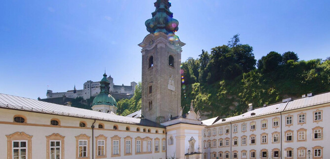 Historická architektura s vysokou věží, obklopená zelenou krajinou. Nebe je jasné a modré, což vytváří klidnou atmosféru. | © Erzabtei St. Peter