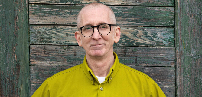 A man with glasses stands in front of a wooden wall. He is wearing a bright yellow shirt and is smiling slightly. | © Robert Peres