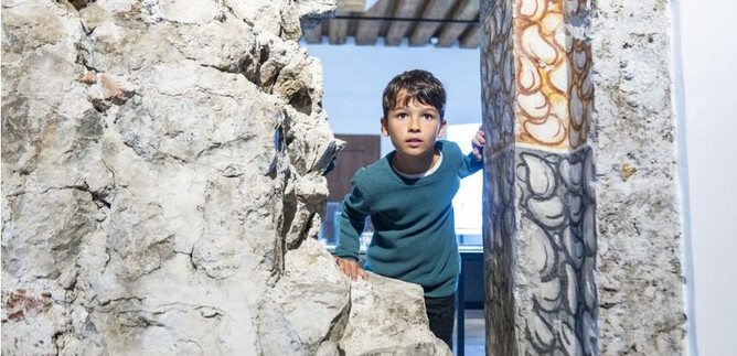 A boy looks curiously through a wall opening. In the background, a bright room can be seen. | © Salzburg Museum