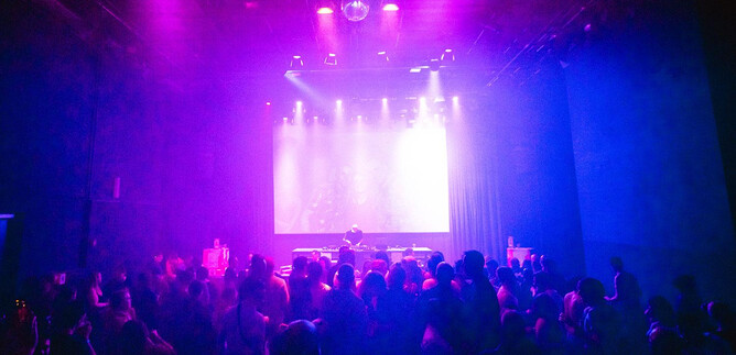 Eine lebhafte Party mit vielen Menschen im Vordergrund. Bunte Lichter und eine DJ-Bühne schaffen eine aufregende Atmosphäre. | © Angelina Widmann