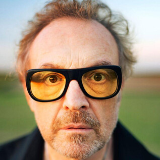 Ein Mann mit Brille und gestyltem Haar blickt direkt in die Kamera. Im Hintergrund ist eine grüne Landschaft zu sehen. | © www.lukasbeck.com