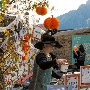 Egy álruhás oroszlán egy világító tököt tart a kezében. Ez egy éjszakai jelenet gyenge fényben, fákkal a háttérben. | © Zoo Salzburg Hellbrunn