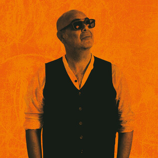 Ein Mann mit Glatze und Sonnenbrille steht vor einem orangefarbenen Hintergrund. Er trägt eine schwarze Weste und schaut nach oben. | © Tobias Koark-Haberl