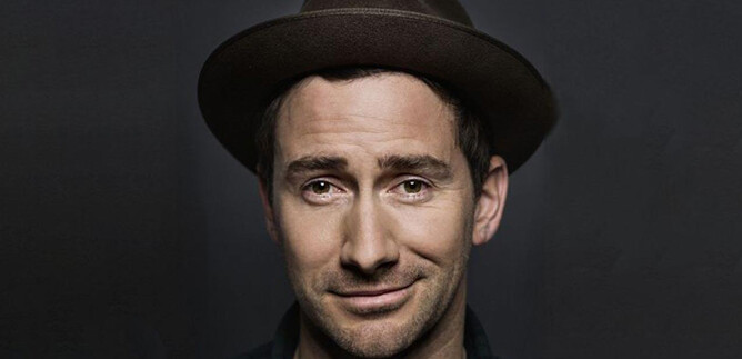 A man with a hat smiles at the camera. The background is dark and plain. | © Christian Brecheis