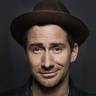 Ein Mann mit einem Hut lächelt in die Kamera. Der Hintergrund ist dunkel und schlicht. | © Christian Brecheis