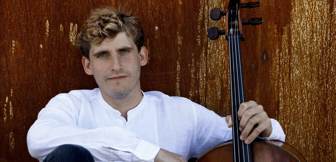 A young man sits relaxed next to a cello. He is wearing a white shirt and is smiling slightly in front of a rustic background wall. | © Ida Wang