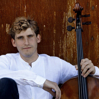 Ein junger Mann sitzt entspannt neben einem Violoncello. Er trägt ein weißes Hemd und lächelt leicht vor einer rustikalen Hintergrundwand. | © Ida Wang