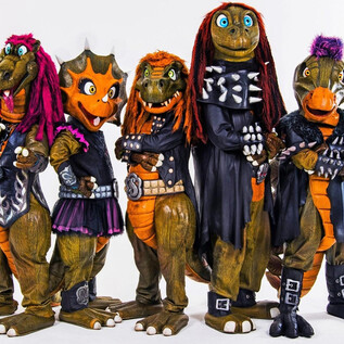 Eine Gruppe von Dinosaurier-Maskottchen in rockigen Outfits steht vor einer dramatischen Hintergrundlandschaft. Der Text "Heavy Saurus" ist prominently platziert. | © sonymusic.de