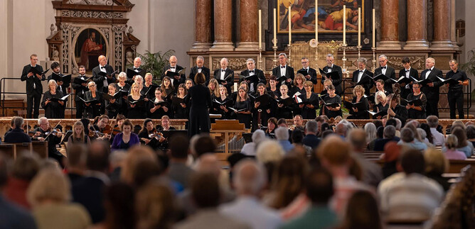 Un coro está en un escenario en una iglesia y canta. En el fondo se pueden ver el altar y numerosos espectadores. | © Dom zu Salzburg