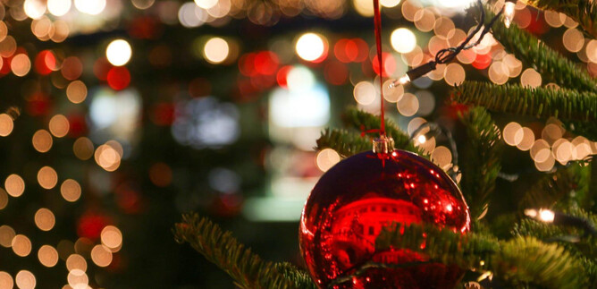 A red Christmas tree ornament hangs on a tree. Bright lights and Christmas decorations shine in the background. | © Hellbrunner Adventzauber