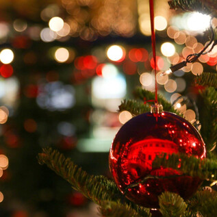 Ein roter Weihnachtsbaum-Schmuck hängt an einem Baum. Im Hintergrund leuchten helle Lichter und Weihnachtsdekorationen. | © Hellbrunner Adventzauber