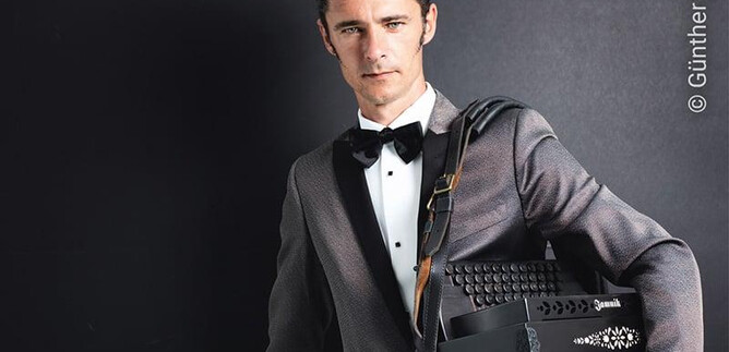 A well-dressed man is holding a black accordion and standing in front of a neutral background. He is wearing a tuxedo with a bow tie and has a confident expression. | © Günther Egger