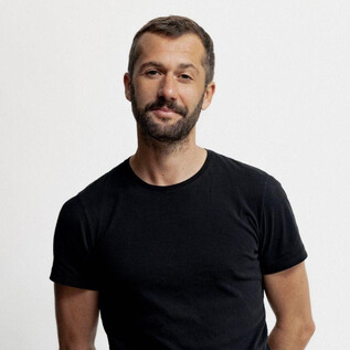 Un homme se tient devant un fond neutre. Il porte un T-shirt noir et a une légère barbe. | © Susan Buth