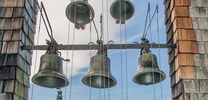 A series of bells hangs on a framework, surrounded by a beautiful sky. In the background, a tower with a green dome is visible. | © SalzburgerLand Tourismus / Eva trifft