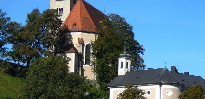 Eine Kirche mit einem hohen Turm und einem roten Dach steht auf einem Hügel. Darunter befinden sich einige Bäume und ein kleines Gebäude. | © Pfarre Mülln