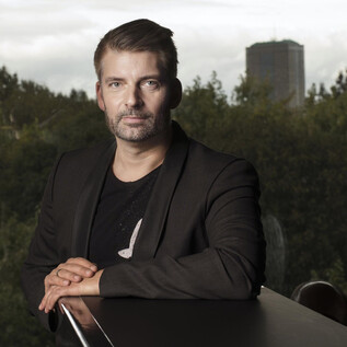 A man in a black blazer is sitting at a table and looking directly at the camera. In the background, trees and a high-rise building are visible. | © Franck Ferville