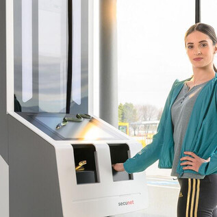 A woman stands at modern self-service terminals at the airport. In the background, there are windows overlooking the airport operations. | © SLT / Christian Krautzberger