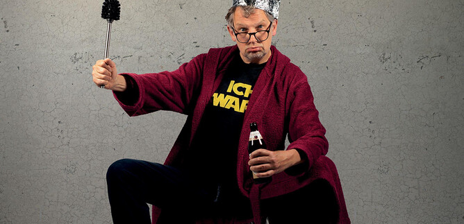 A man in a red bathrobe is wearing an aluminum hat and holding a bottle of beer in one hand and a broom in the other. He is sitting with a confident expression in front of a gray background. | © Dominic Reichenbach