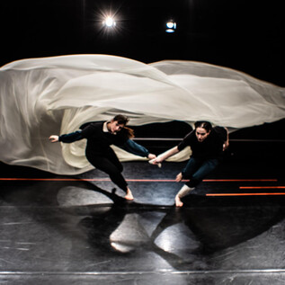 Zwei Tänzerinnen bewegen sich dynamisch über eine Dunkle Fläche, während ein leichter Stoff über ihnen schwingt. Das Bild strahlt Energie und Bewegung aus. | © Fabian Schober