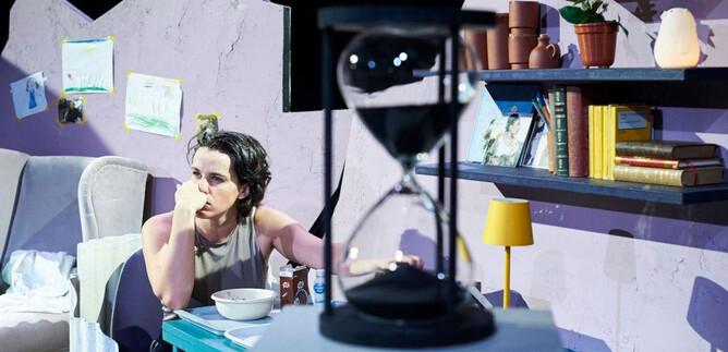 Eine Person sitzt nachdenklich an einem Tisch in einem schlicht eingerichteten Raum. Hinter ihr stehen Bücherregale und eine Sanduhr ist im Vordergrund zu sehen. | © Tom Leather Neumeier