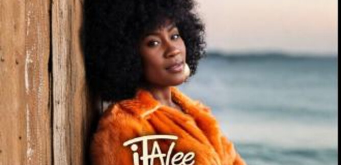 Eine Frau mit lockigem Haar trägt einen orangefarbenen Mantel und lehnt an einer Holzwand. Im Hintergrund ist das Meer und der Himmel zu sehen. | © Jazzit