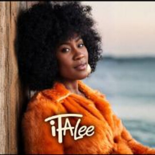 Une femme aux cheveux bouclés porte un manteau orange et s'appuie contre un mur en bois. À l'arrière-plan, on peut voir la mer et le ciel. | © Jazzit