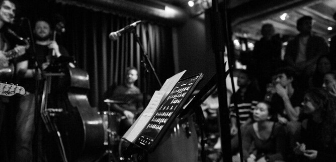 Élő zenei előadás egy kis, hangulatos helyszínen. A zenészek a színpadon játszanak, miközben a közönség figyelmesen nézi őket. | © Jazzit