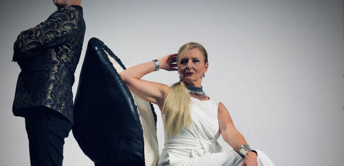 Ein Mann und eine Frau posieren vor einem schlichten Hintergrund. Die Frau sitzt auf einem übergroßen Schuh und trägt ein elegantes weißes Outfit. | © Beatriz Teixeira