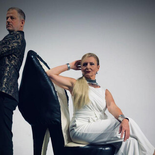 Ein Mann und eine Frau posieren vor einem schlichten Hintergrund. Die Frau sitzt auf einem übergroßen Schuh und trägt ein elegantes weißes Outfit. | © Beatriz Teixeira