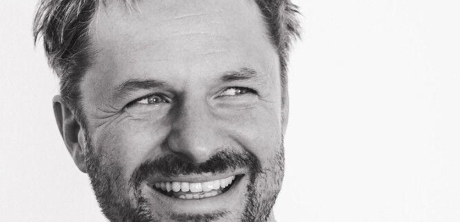 A smiling man with a beard and curly hair. He is wearing a light shirt and radiates joy. | © Stephan Brueckler