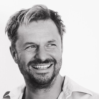 Uśmiechnięty mężczyzna z brodą i kręconymi włosami. Ma na sobie jasną koszulę i emanuje radością. | © Stephan Brueckler