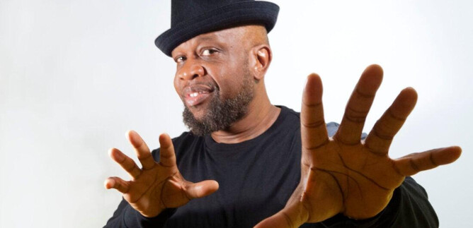 Ein Mann in einem schwarzen Shirt und einem Hut macht mit seinen Händen eine auffällige Geste. Er hat ein freundliches Lächeln auf dem Gesicht. | © Jazzit