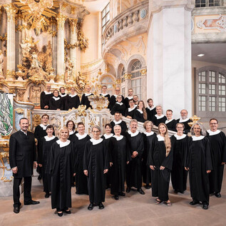 A group of choir members stands outdoors in front of a blooming bush. They are wearing black clothing and colorful scarves. | © Tobias Ritz