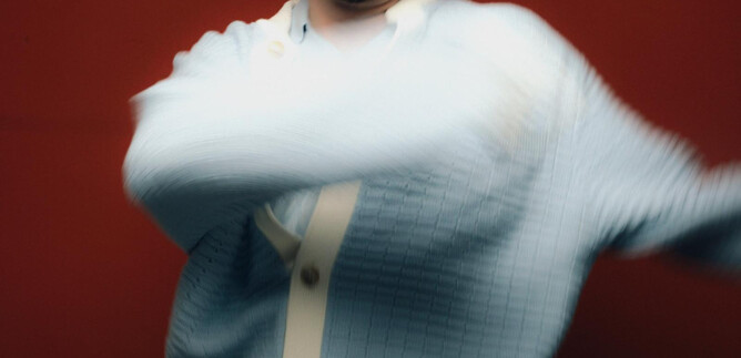 A young man in a light blue sweater poses against a red background. He moves with a relaxed posture. | © töchtersöhne.org