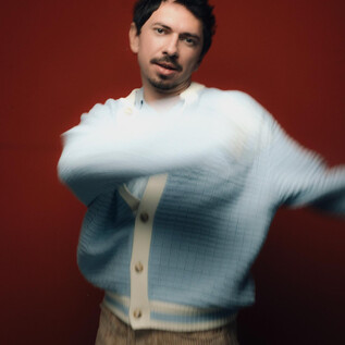 A young man in a light blue sweater poses against a red background. He moves with a relaxed posture. | © töchtersöhne.org