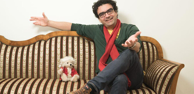 Un uomo è seduto rilassato su un divano e sorride. Accanto a lui c'è un piccolo cane con una sciarpa rossa. | © Wolfgang Lienbacher