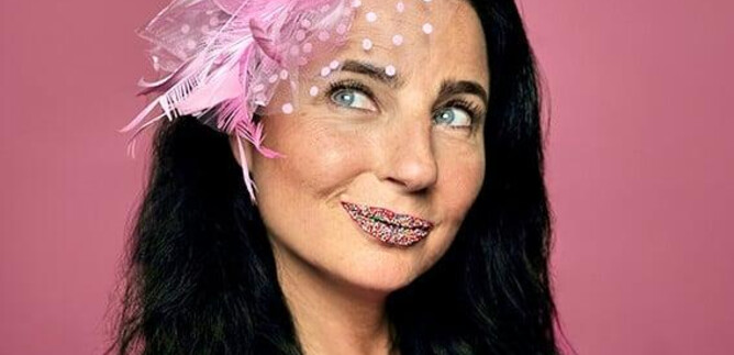 A woman with black hair is wearing a striking pink headpiece. Her face is adorned with glittering lipstick, and she playfully smiles at the camera. | © Sabine Stieger