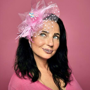 Eine Frau mit schwarzem Haar trägt einen auffälligen rosa Kopfschmuck. Ihr Gesicht ist mit glitzerndem Lippenstift geschmückt und sie lächelt spielerisch in die Kamera. | © Sabine Stieger