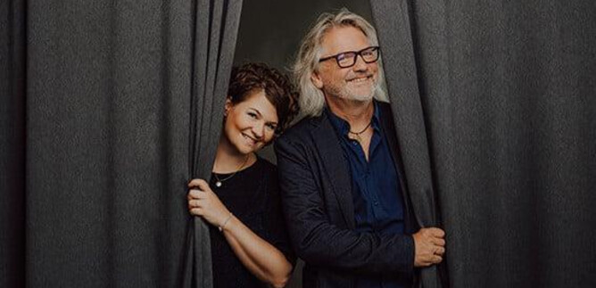 Pár se šťastně dívá za závěsem. Oba se usmívají a vypadají přátelsky. | © Hannes Höchsmann