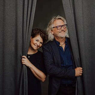 Ein Paar schaut fröhlich hinter einem Vorhang heraus. Beide lächeln und wirken freundlich. | © Hannes Höchsmann