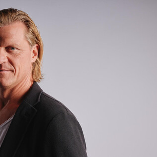 Ein Mann mit längeren, blonden Haaren und einem schwarzen Blazer steht vor einem neutralen Hintergrund. Er blickt direkt in die Kamera und wirkt selbstbewusst. | © Jeff Mangione