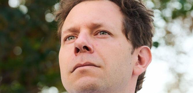 A man with slightly wavy hair looks forward. The background is blurred and shows green plants. | © Kurt Prinz