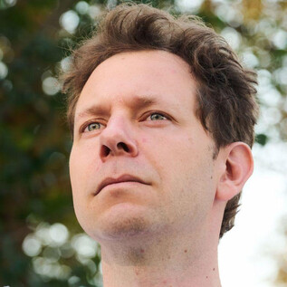 A man with slightly wavy hair looks forward. The background is blurred and shows green plants. | © Kurt Prinz