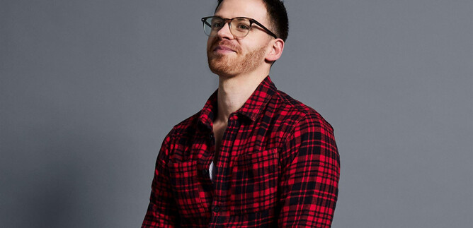 A man is wearing a checked red shirt and glasses. He is smiling and standing in front of a gray background. | © Andreas Kusy